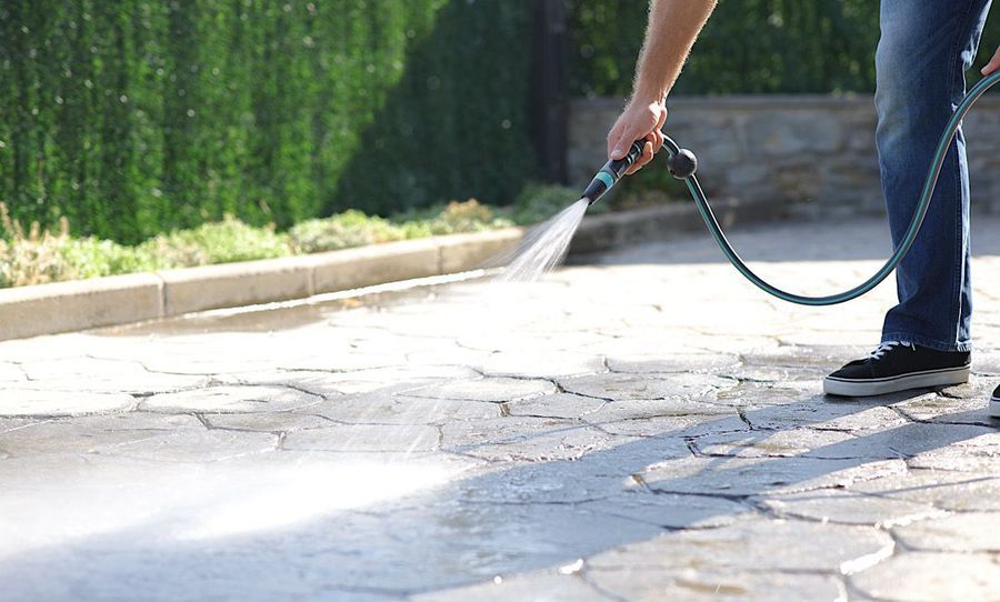 Person spraying water from a hose onto a sunlit paved driveway